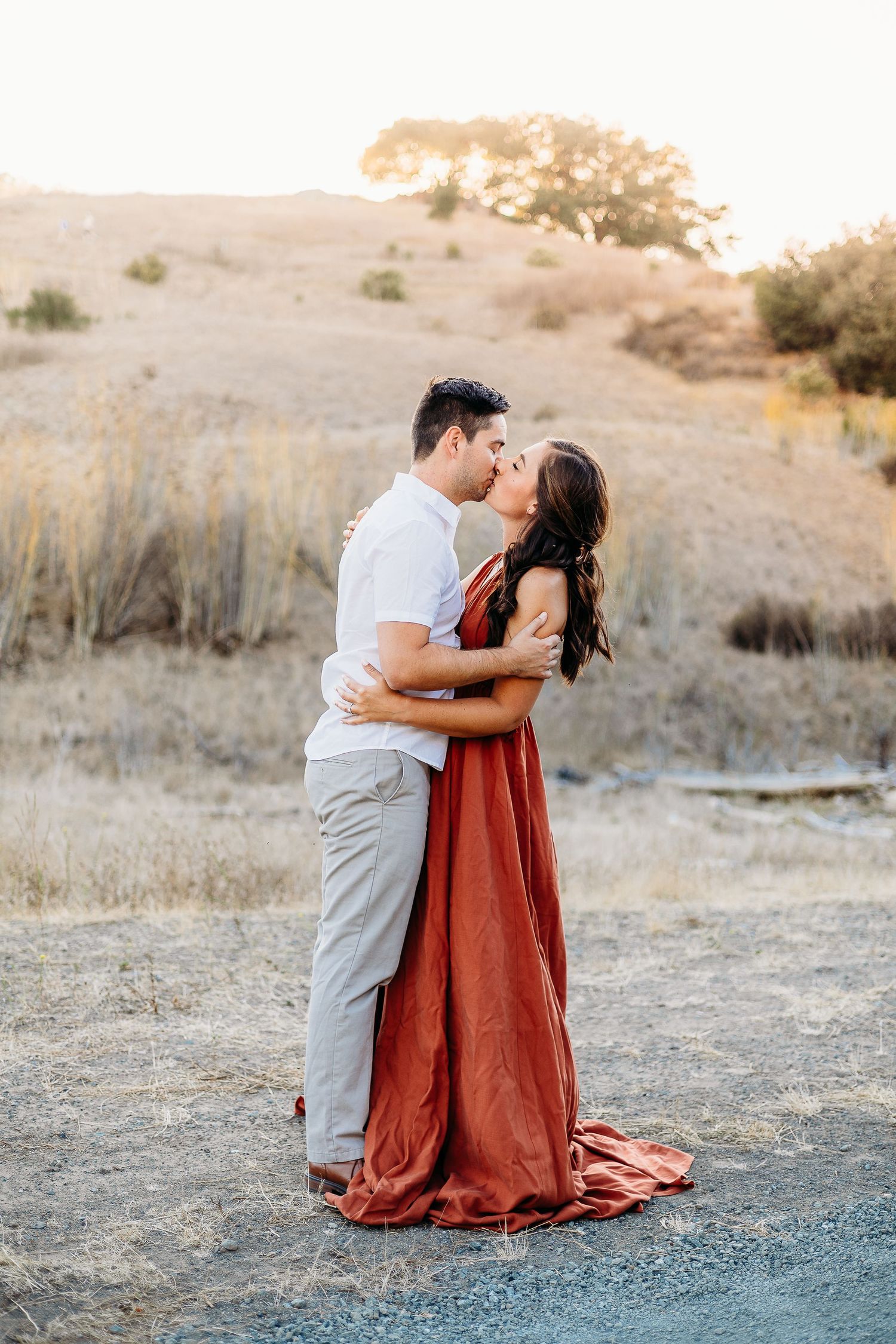 Romantic embrace between couple wearing white and rust colored clothing on hillside path.