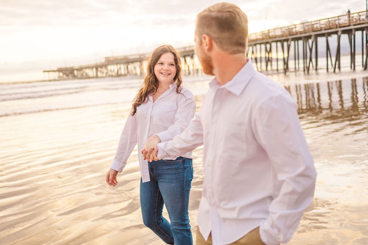 A couple shares a playful moment walking together on a beach with a long pier behind them.