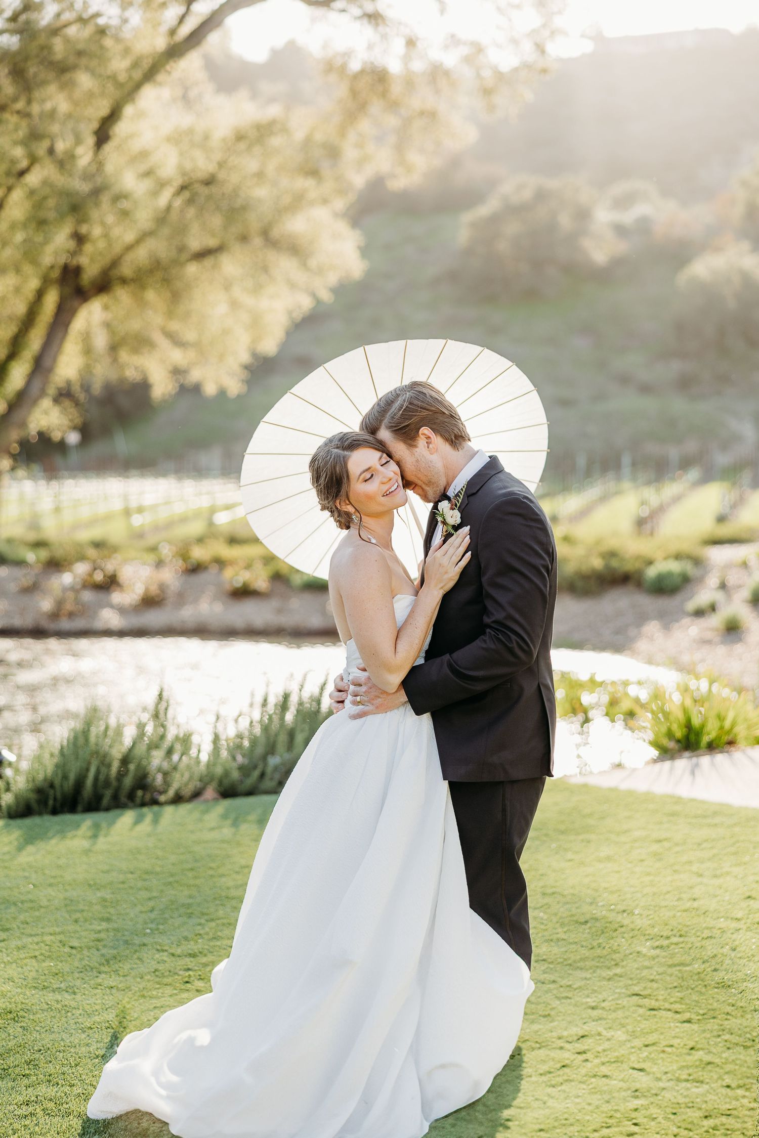 A romantic wedding couple shares a tender kiss under a white parasol in a sunlit garden setting.
