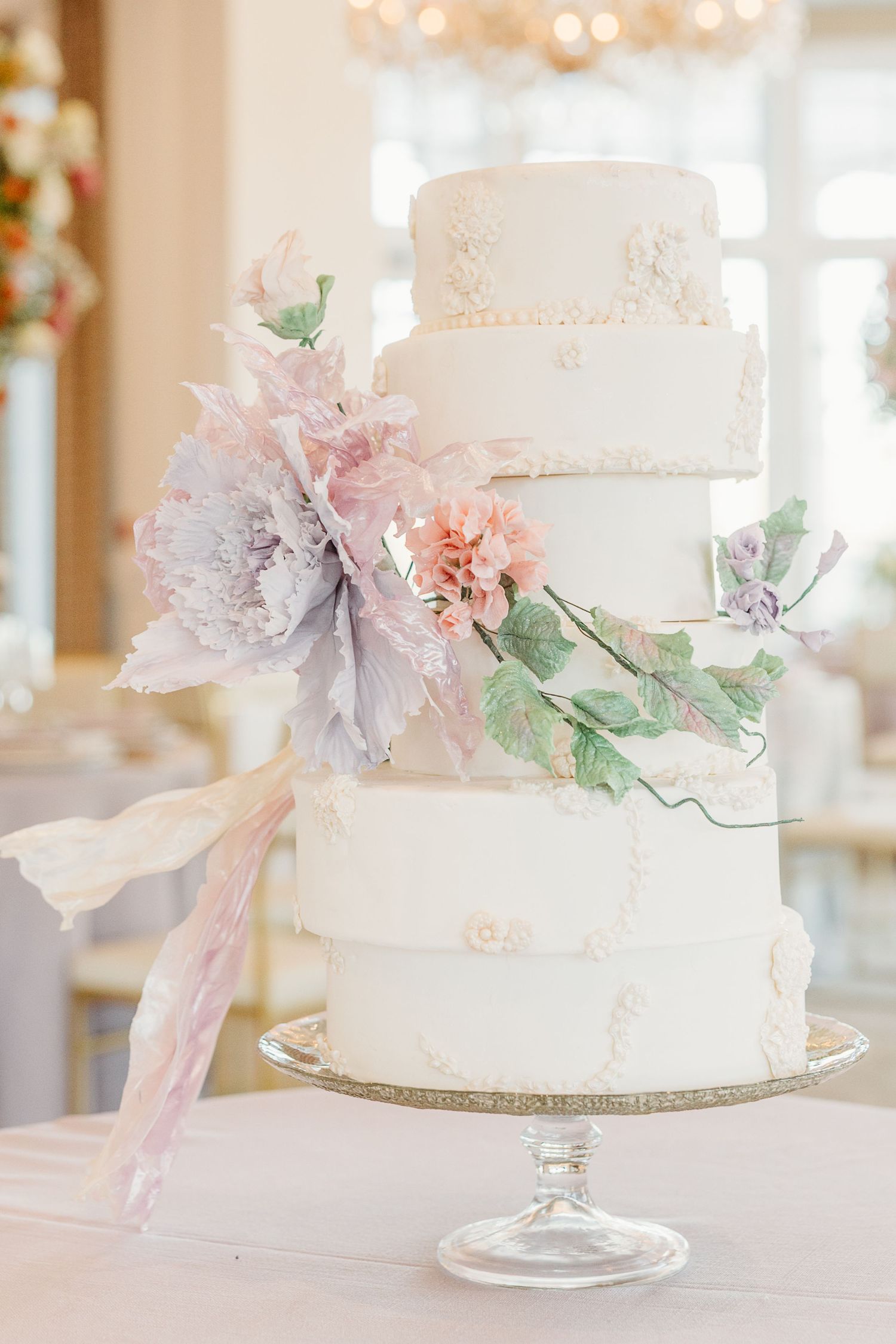 Elegant white wedding cake decorated with delicate pastel pink and lavender sugar flowers on a crystal cake stand.