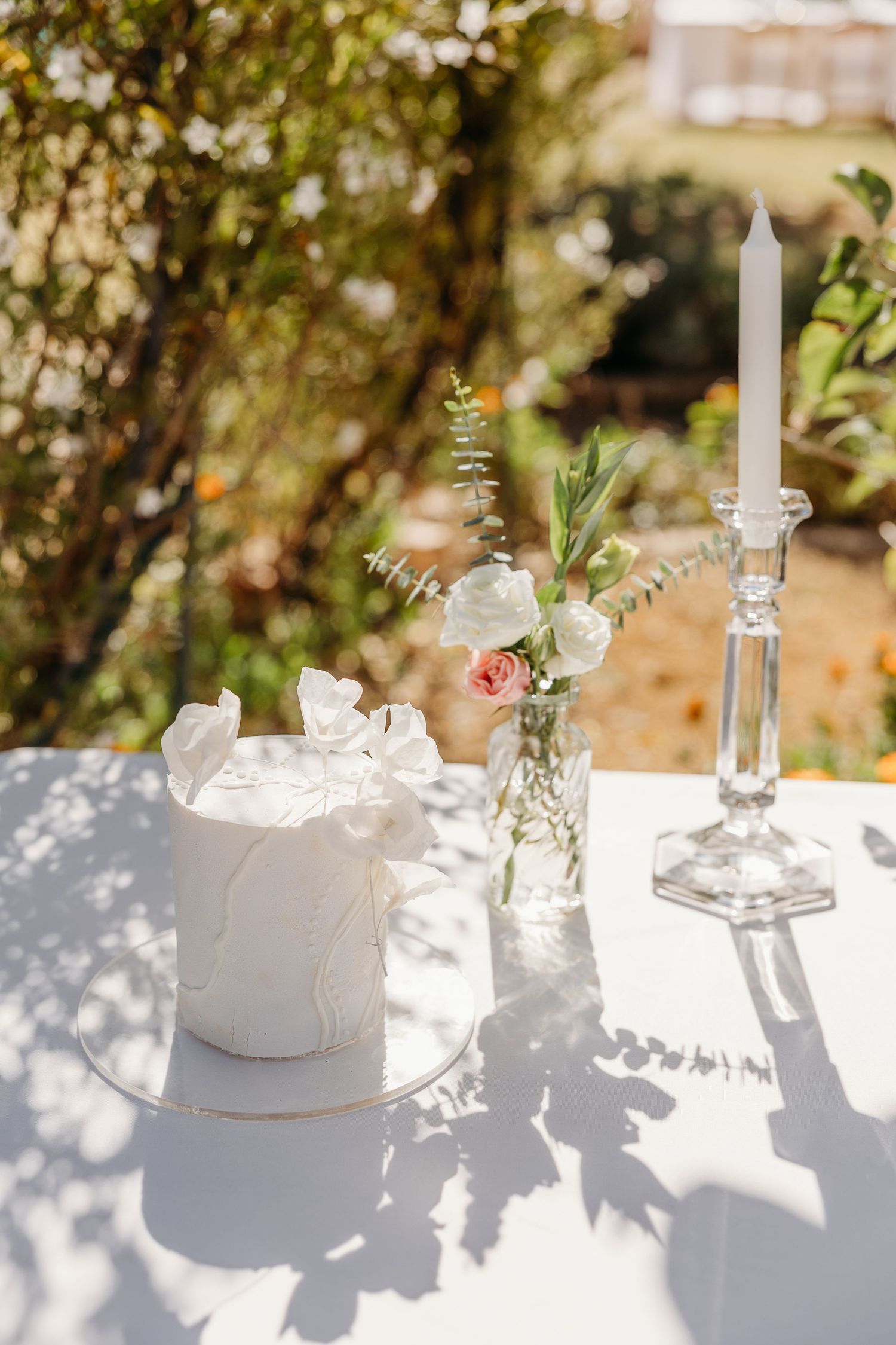 Elegant white wedding cake and floral arrangements on a sunlit table creating dappled shadows.