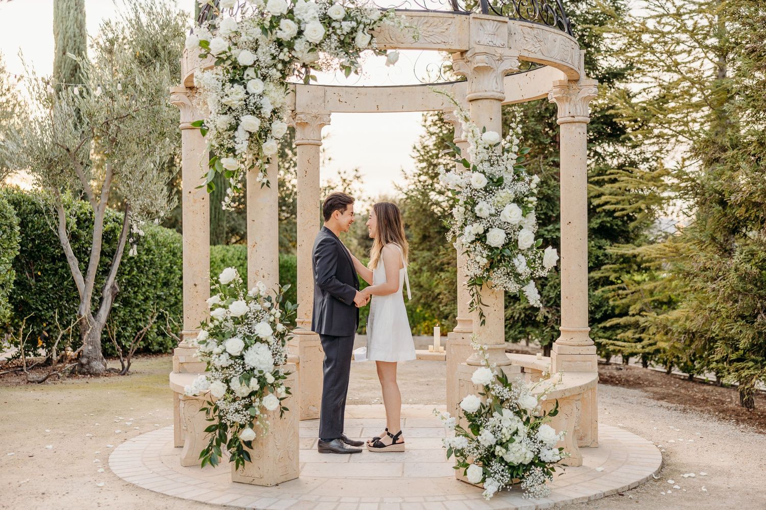 A romantic outdoor wedding ceremony takes place under an elegant stone gazebo decorated with white floral arrangements.