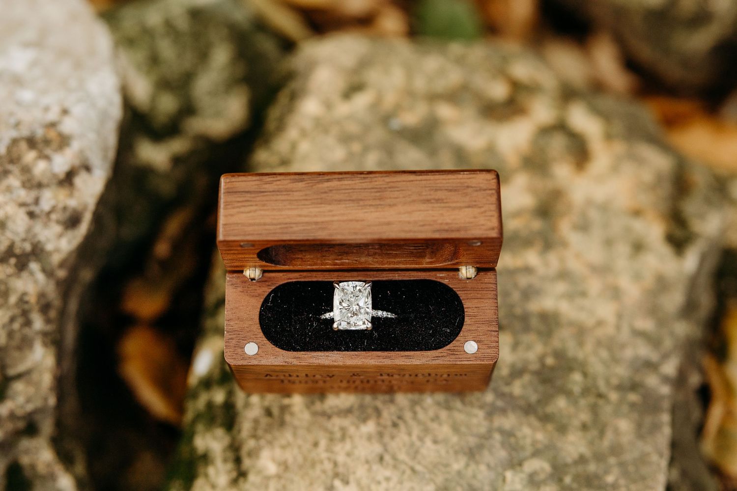 Close-up of wooden ring box with engagement ring displayed on natural stone surface.