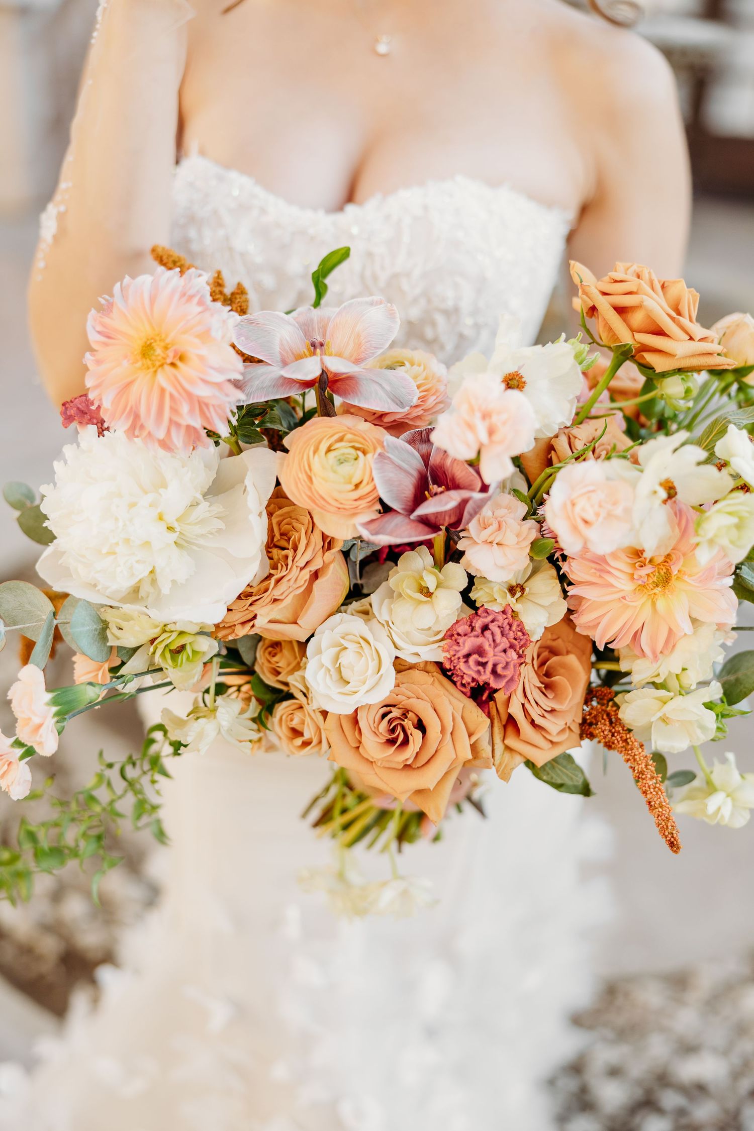 Romantic peach and coral wedding bouquet with roses, dahlias and mixed flowers against a white dress.