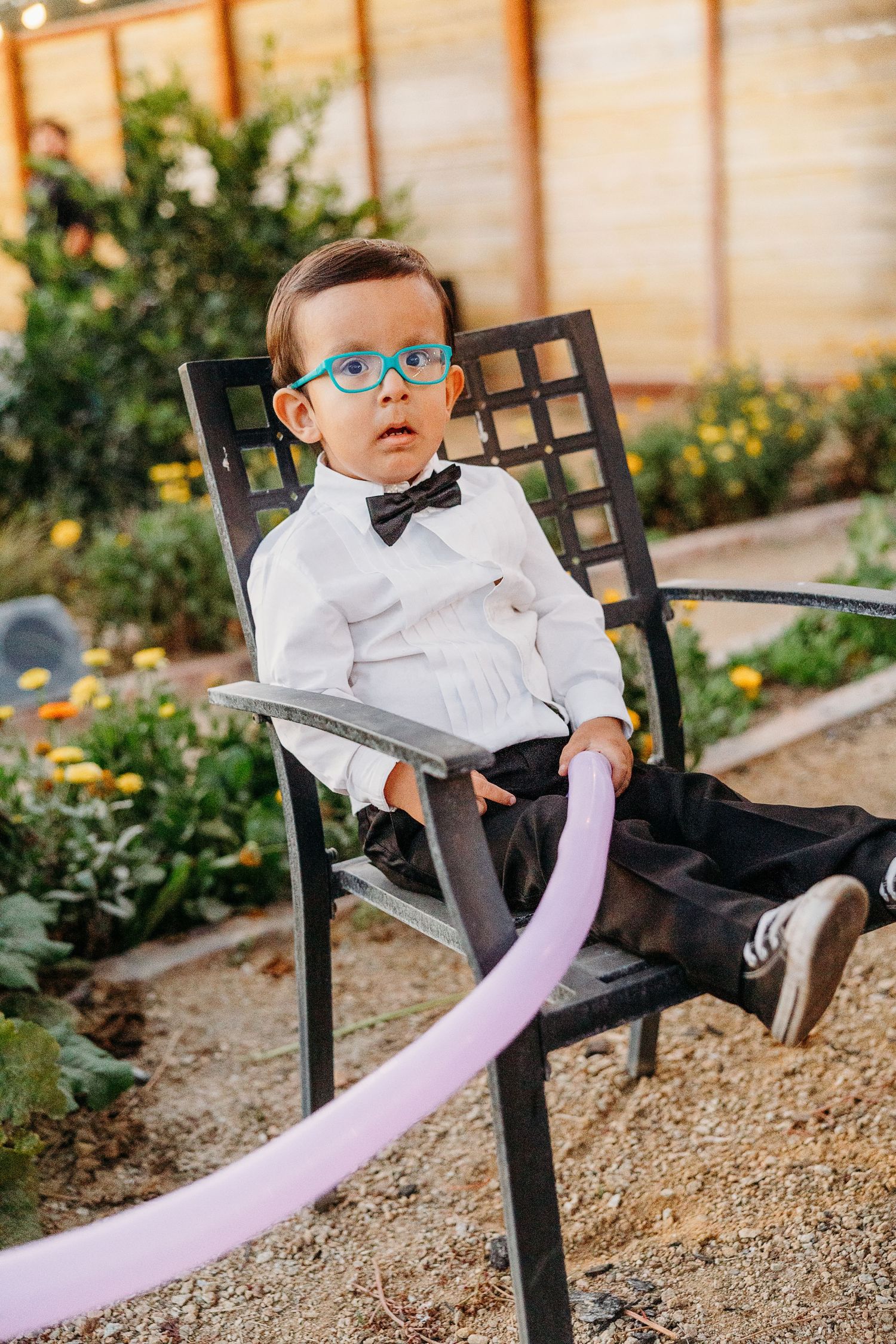 Baby wearing glasses and bowtie lounges in black metal patio chair with pink blanket in garden setting.