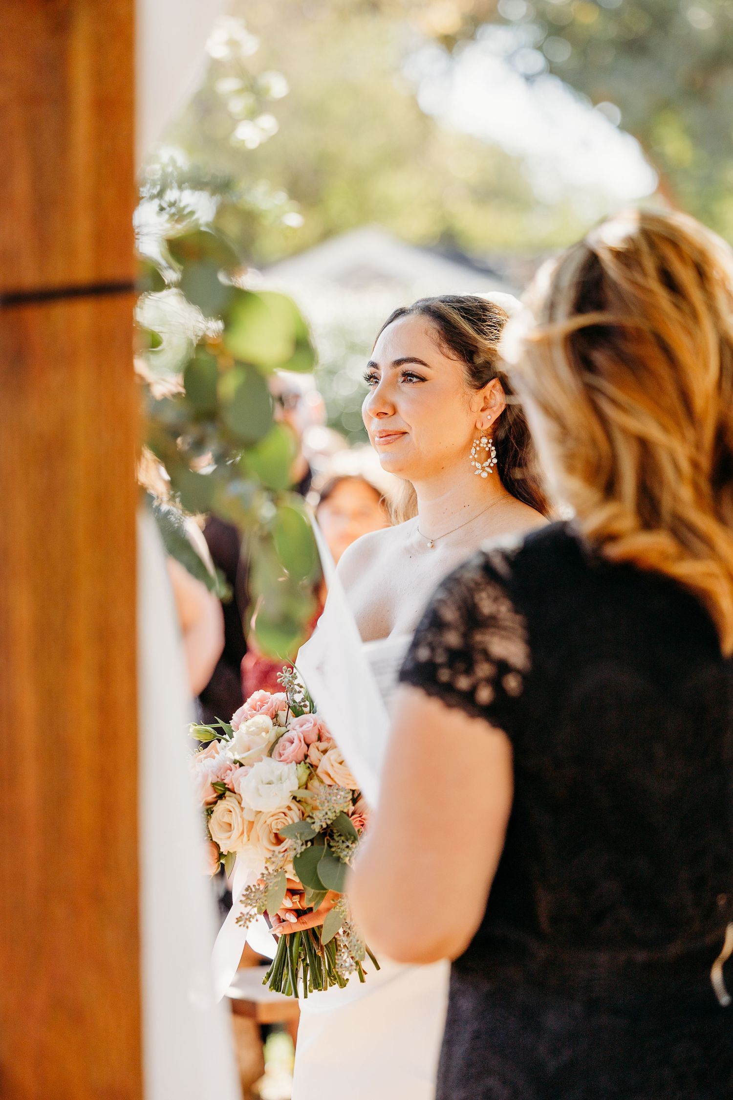 Bridesmaids in black dresses hold bouquets during an outdoor wedding ceremony with wooden beams overhead.