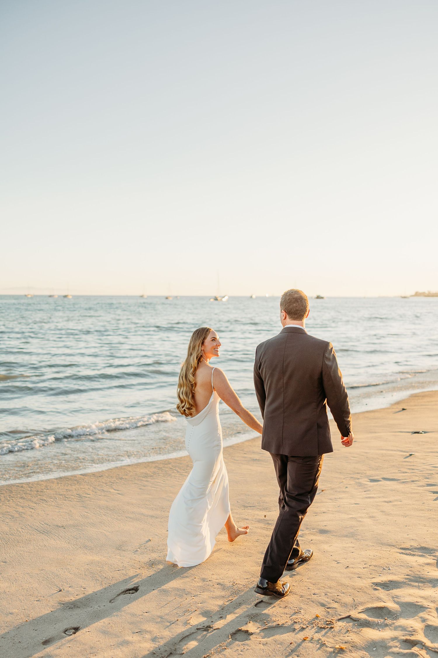 A couple walks hand in hand along a sandy beach at sunset with ocean waves in the background.