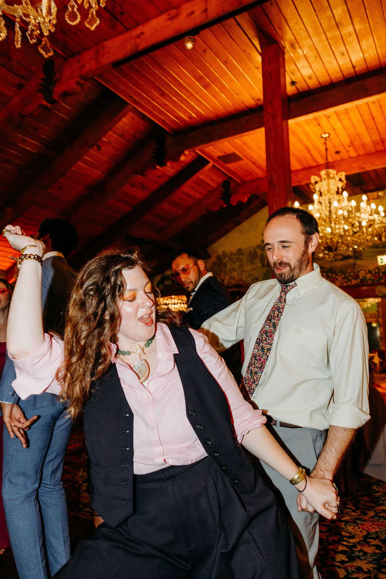 Wedding reception dance party under wooden beams and chandeliers with warm lighting.