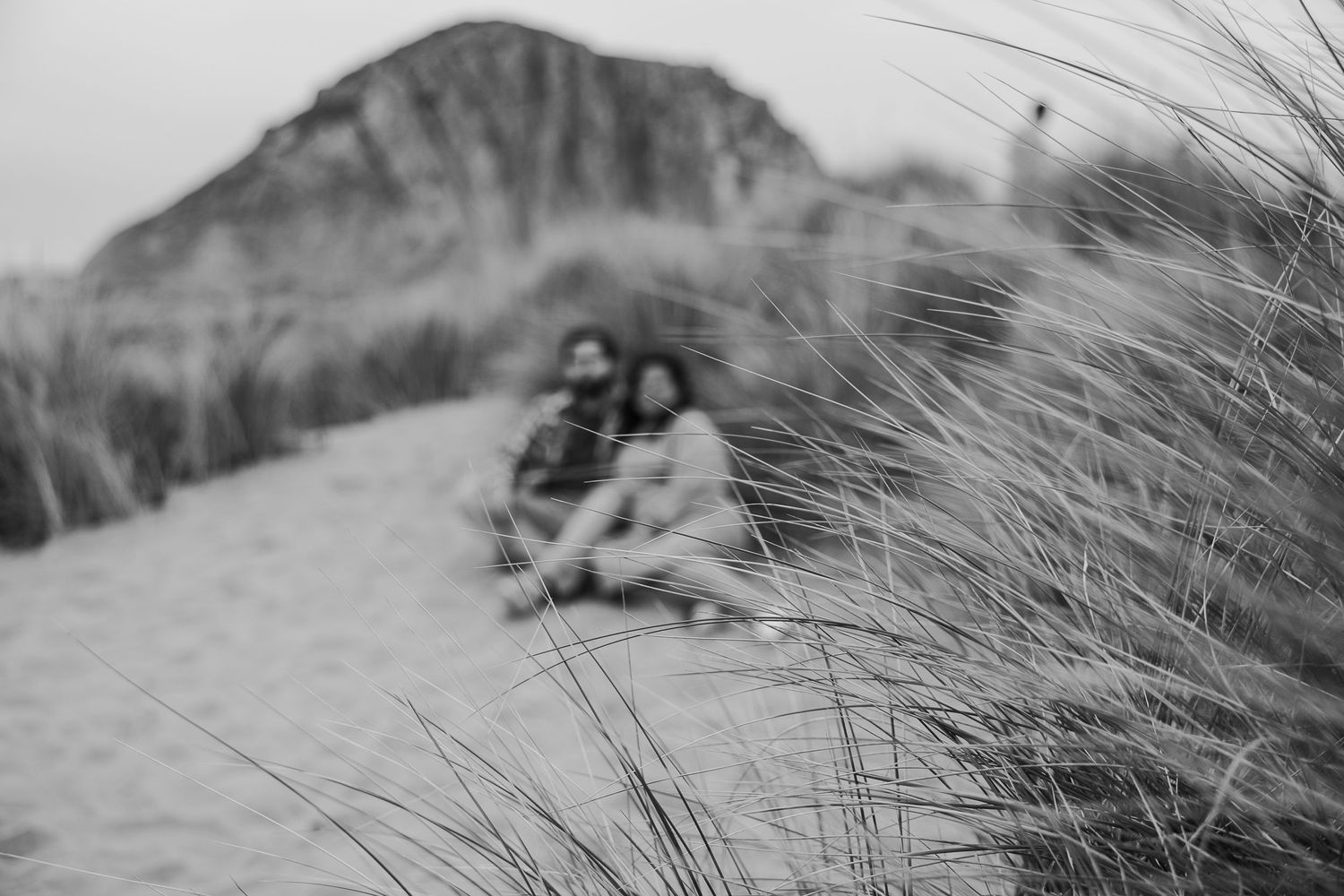 Black and white photo of a couple sitting among beach grass with coastal cliffs in the distance.