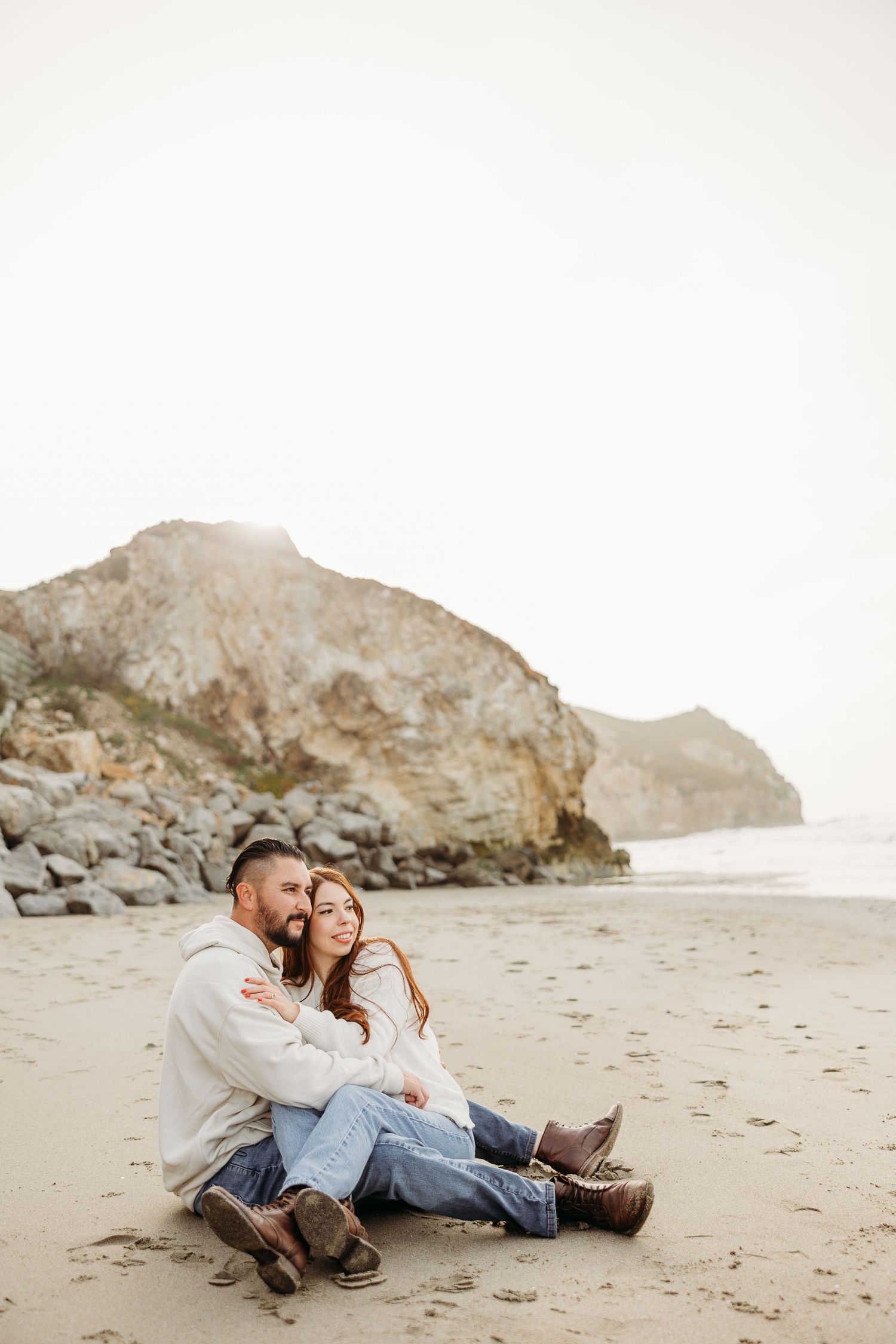 A couple relaxes on a sandy beach with rocky cliffs in the background on a misty overcast day.