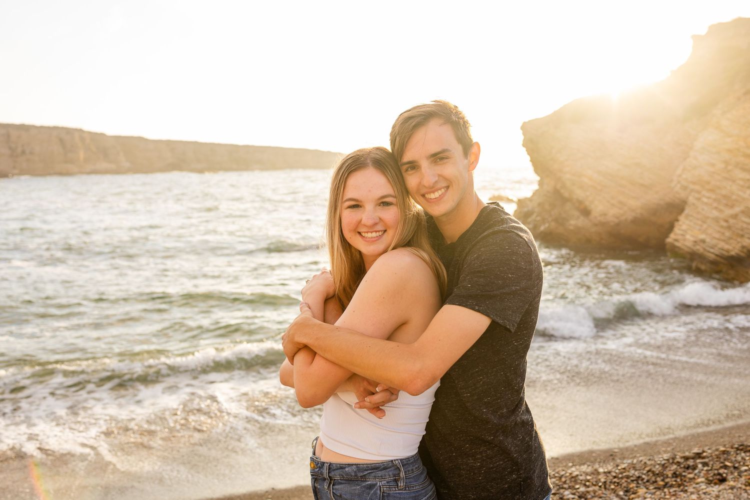 A romantic couple embraces on a beach at sunset with rocky cliffs in the background.