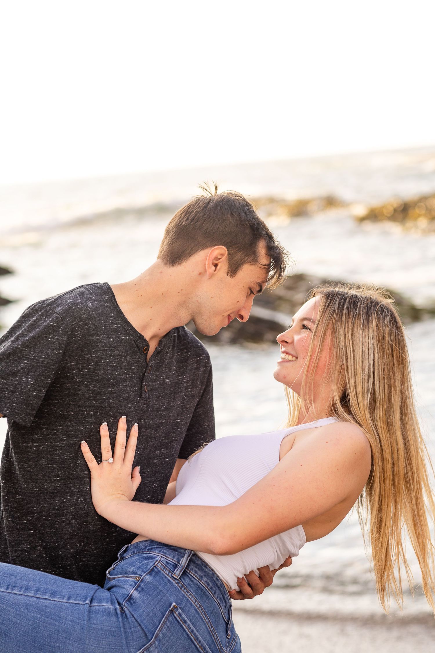 A couple laughs together while embracing on a beach with waves behind them.