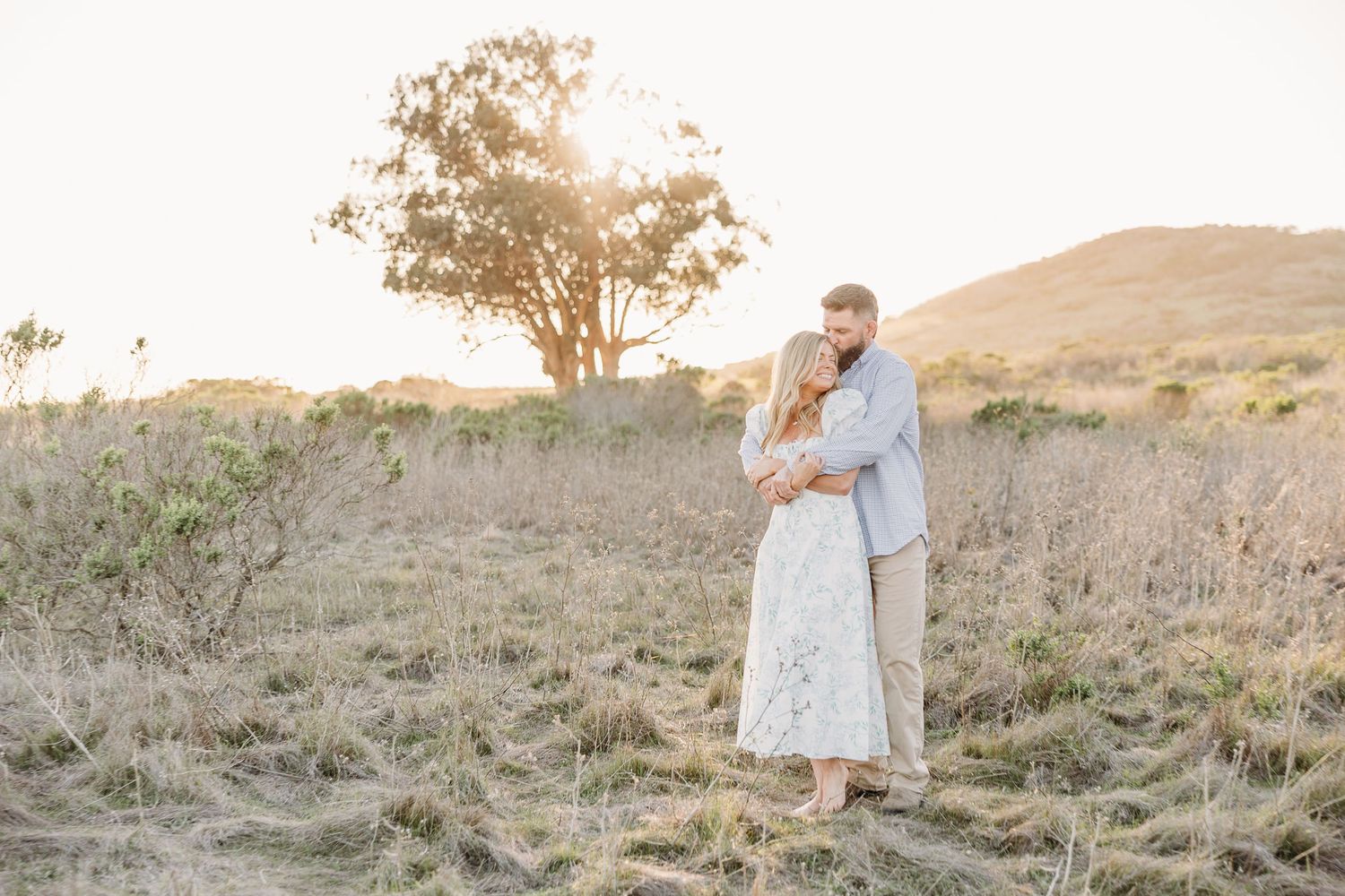 A couple embraces joyfully in front of large coastal rock formations on a sandy beach.