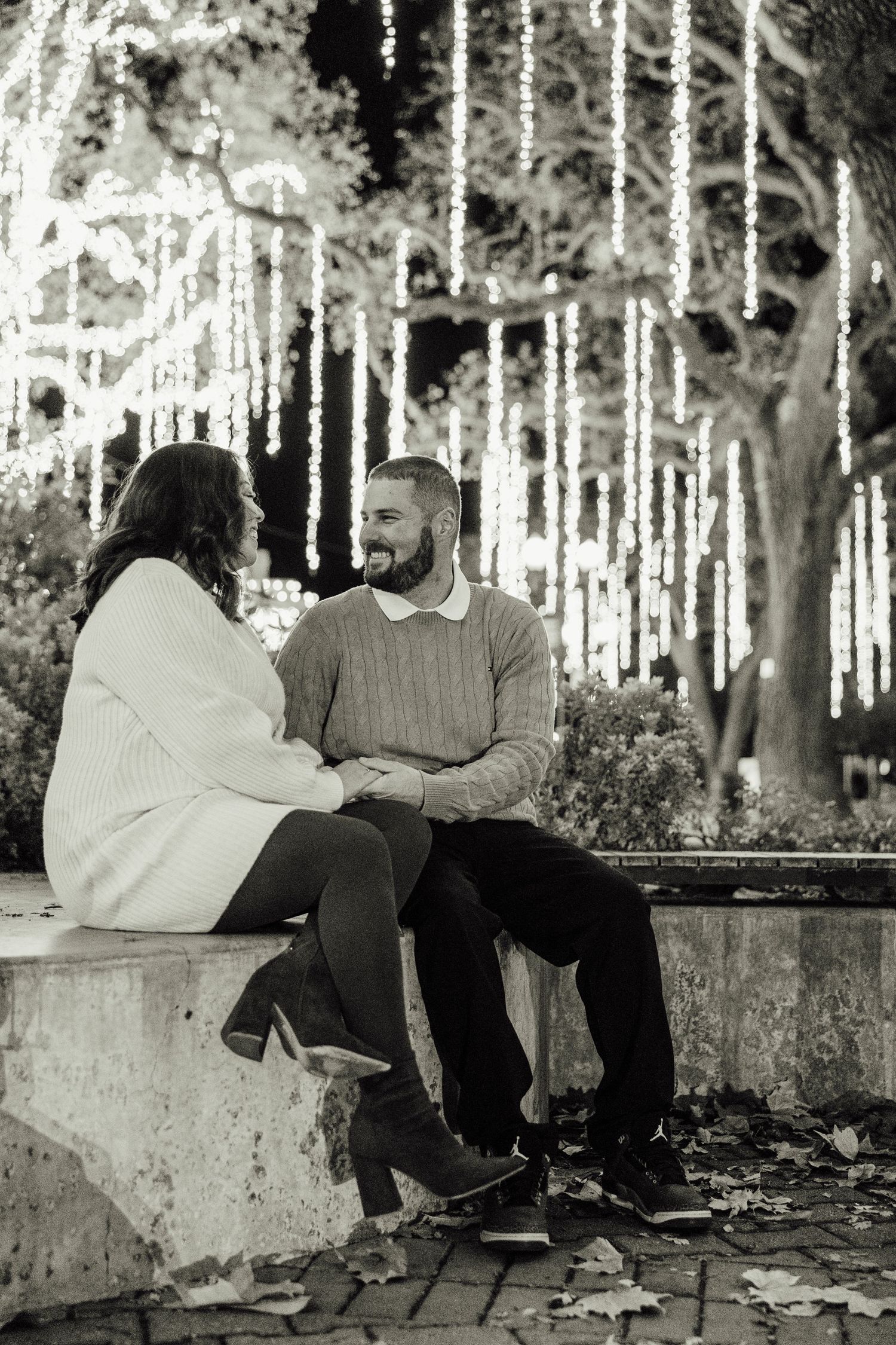 Couple sitting together on a stone ledge surrounded by hanging lights in a dreamy outdoor setting.