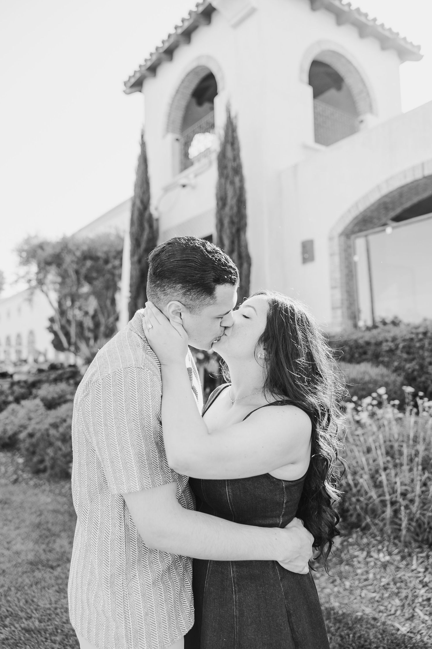 A black and white romantic portrait of a couple kissing outside a Spanish-style building with cypress trees.