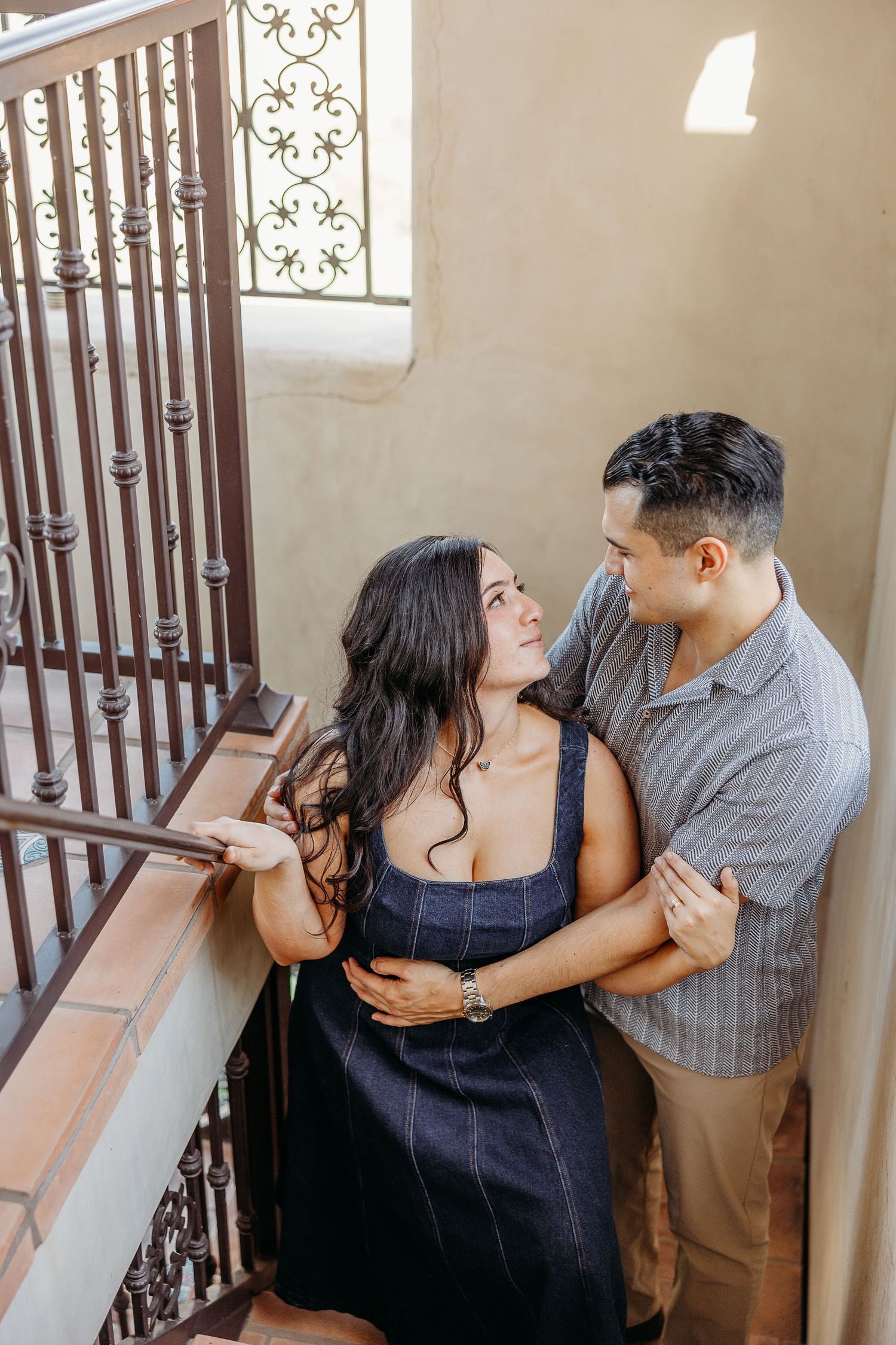 A romantic couple embracing on vintage stairs with ornate metalwork and sunlit walls.