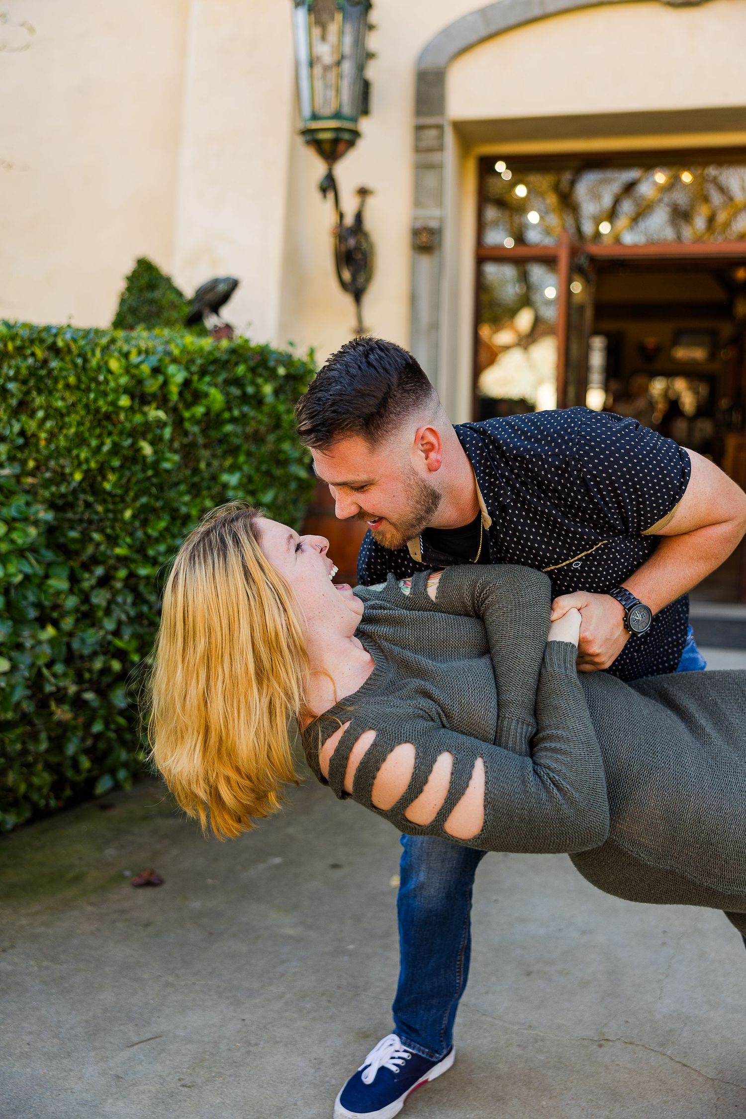 Playful dip pose captured in front of rustic building entrance with lush greenery.