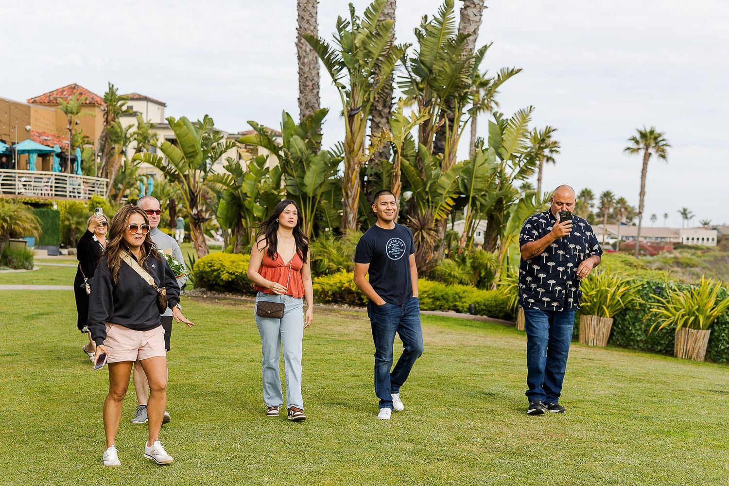 A group of people walking and socializing on a lush green lawn with palm trees and buildings in the background.