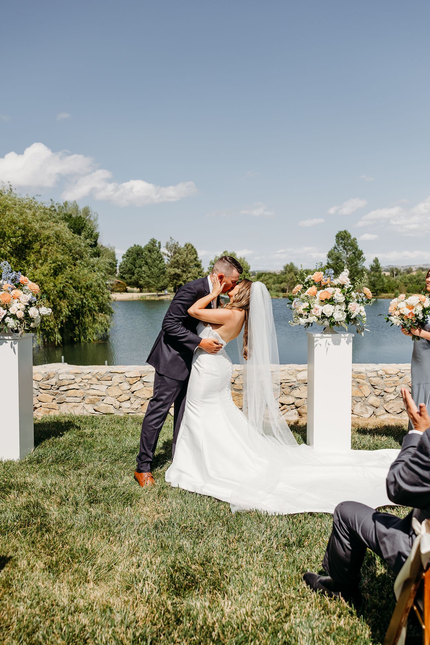 Wedding couple sharing intimate moments by white pillars with floral arrangements against a lakeside backdrop.