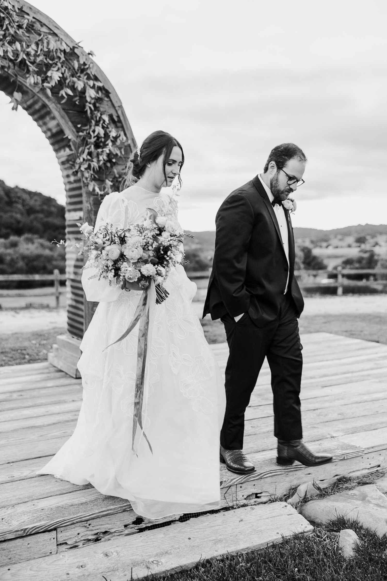 Black and white photo of newlyweds walking on wooden platform under decorative brick archway.