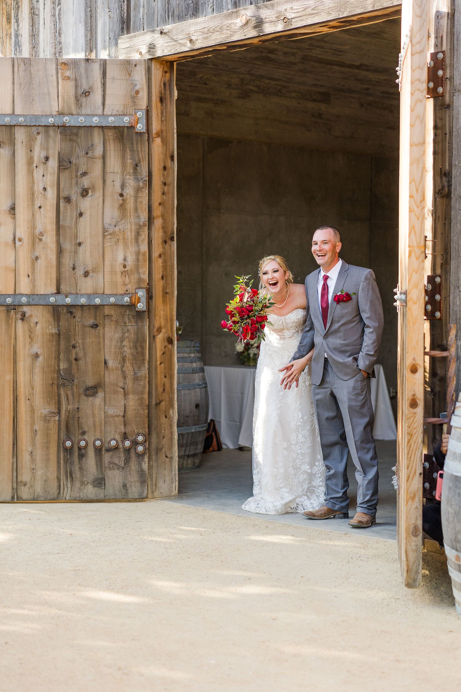 A wedding couple standing in a rustic barn doorway, the bride holding a vibrant red bouquet.