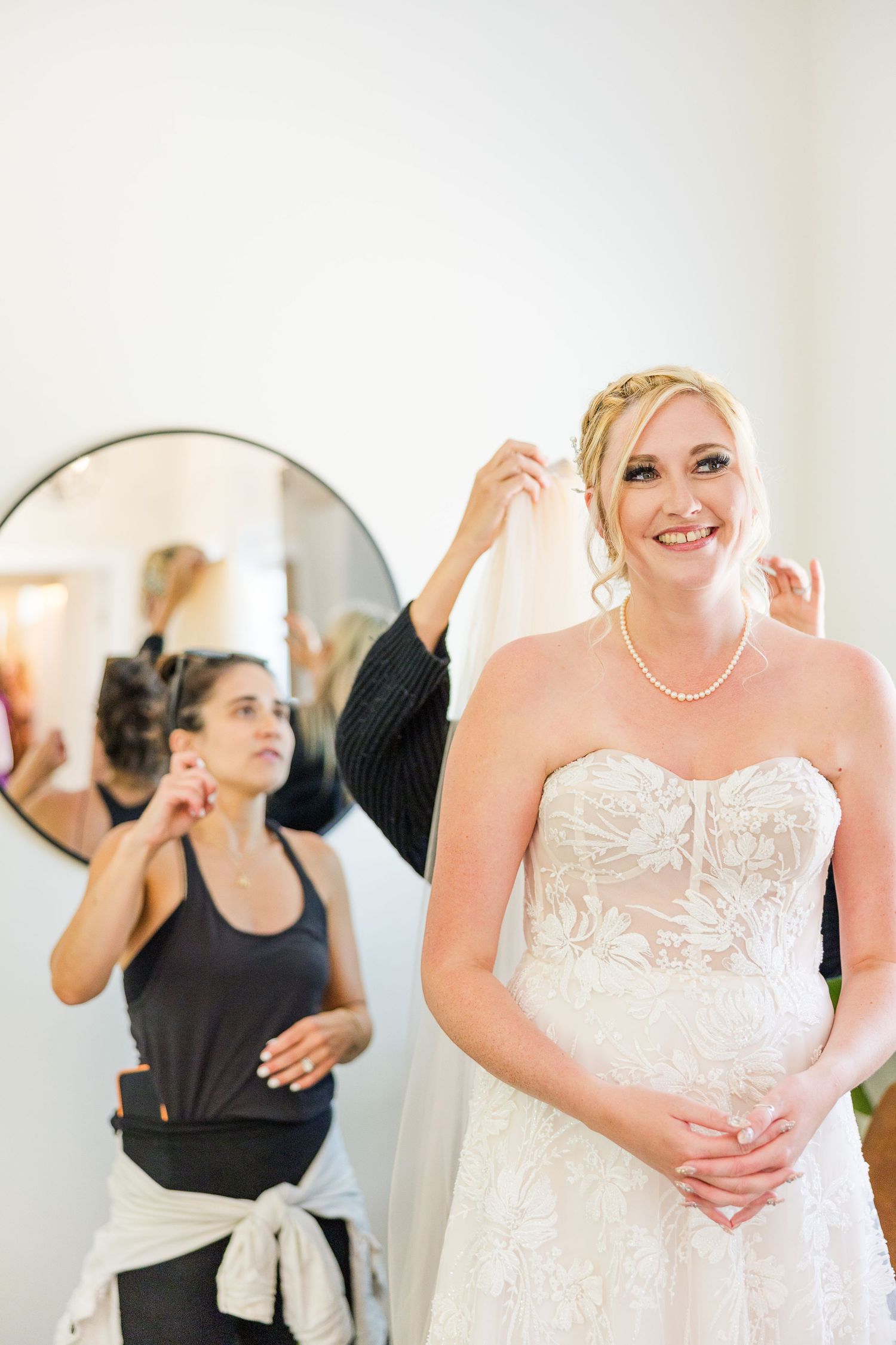 Final wedding preparations with bride in strapless gown getting styled in bright room with round mirror.