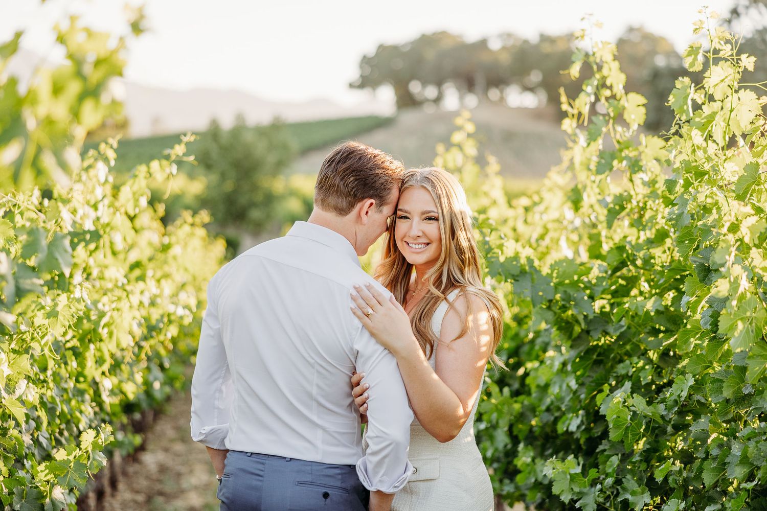 A romantic couple embraces in a sun-drenched vineyard during golden hour with rolling hills in the background.