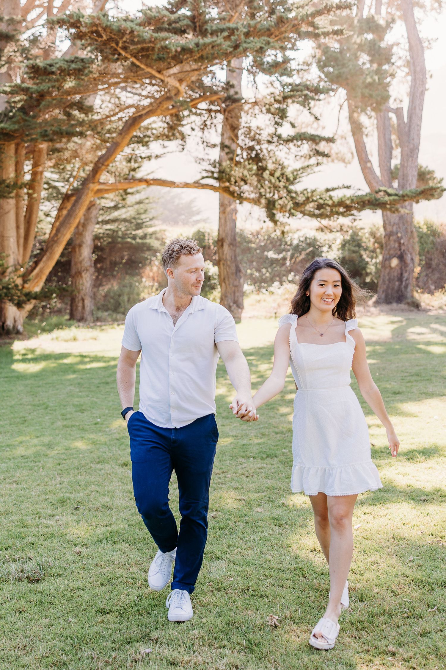 Couple holding hands while walking through park with pine trees in background.