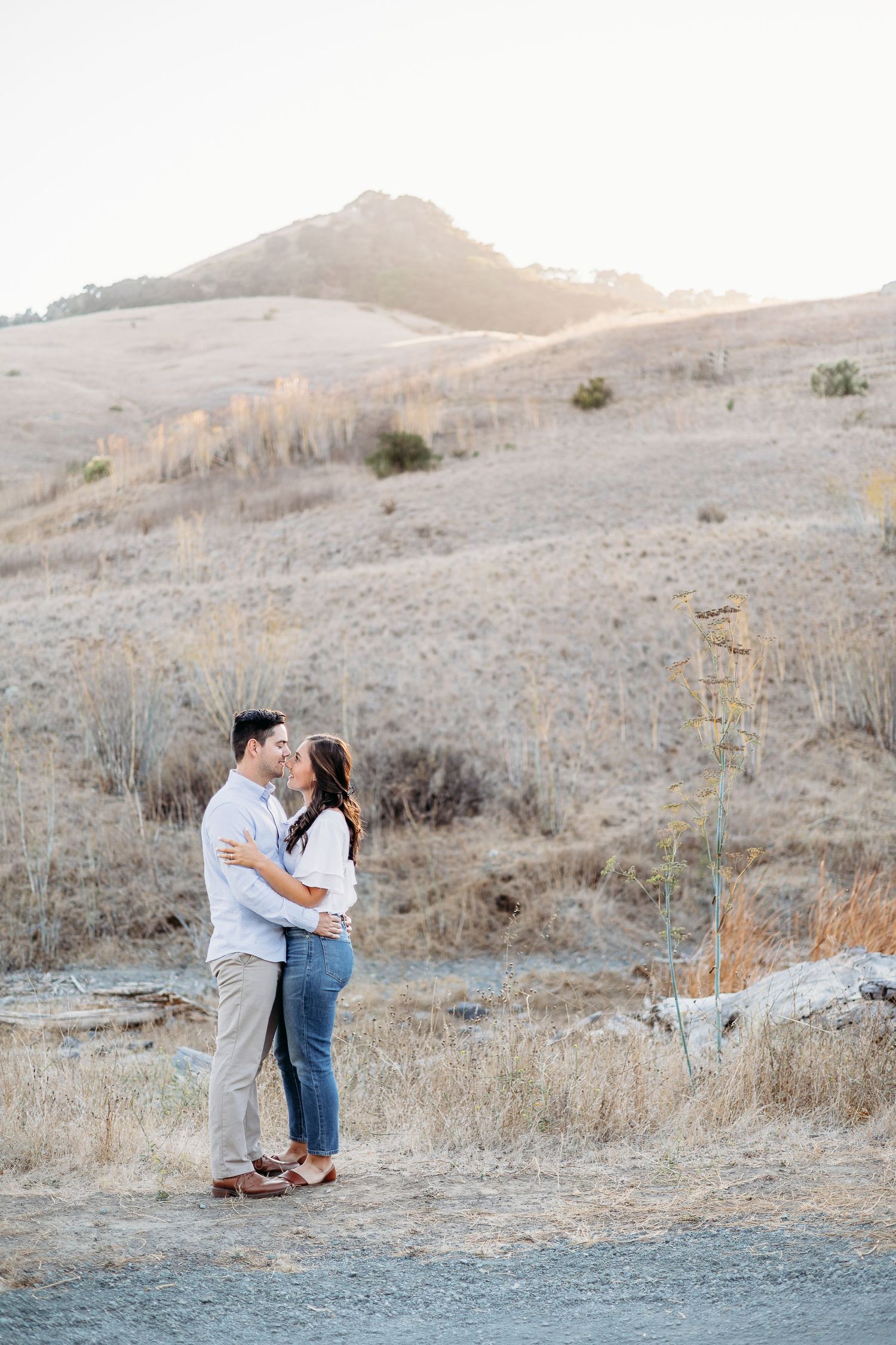 A couple embraces on a hilltop trail during golden hour with mountains in the background.