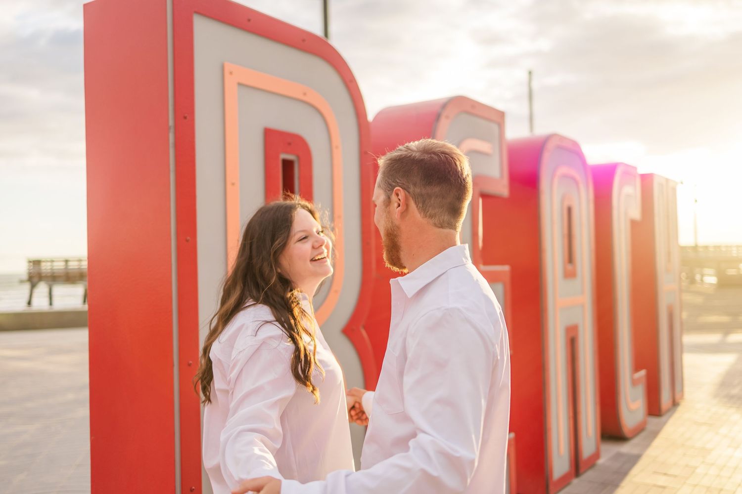 A couple in white shirts poses romantically against large red letters during a warm sunset photoshoot.