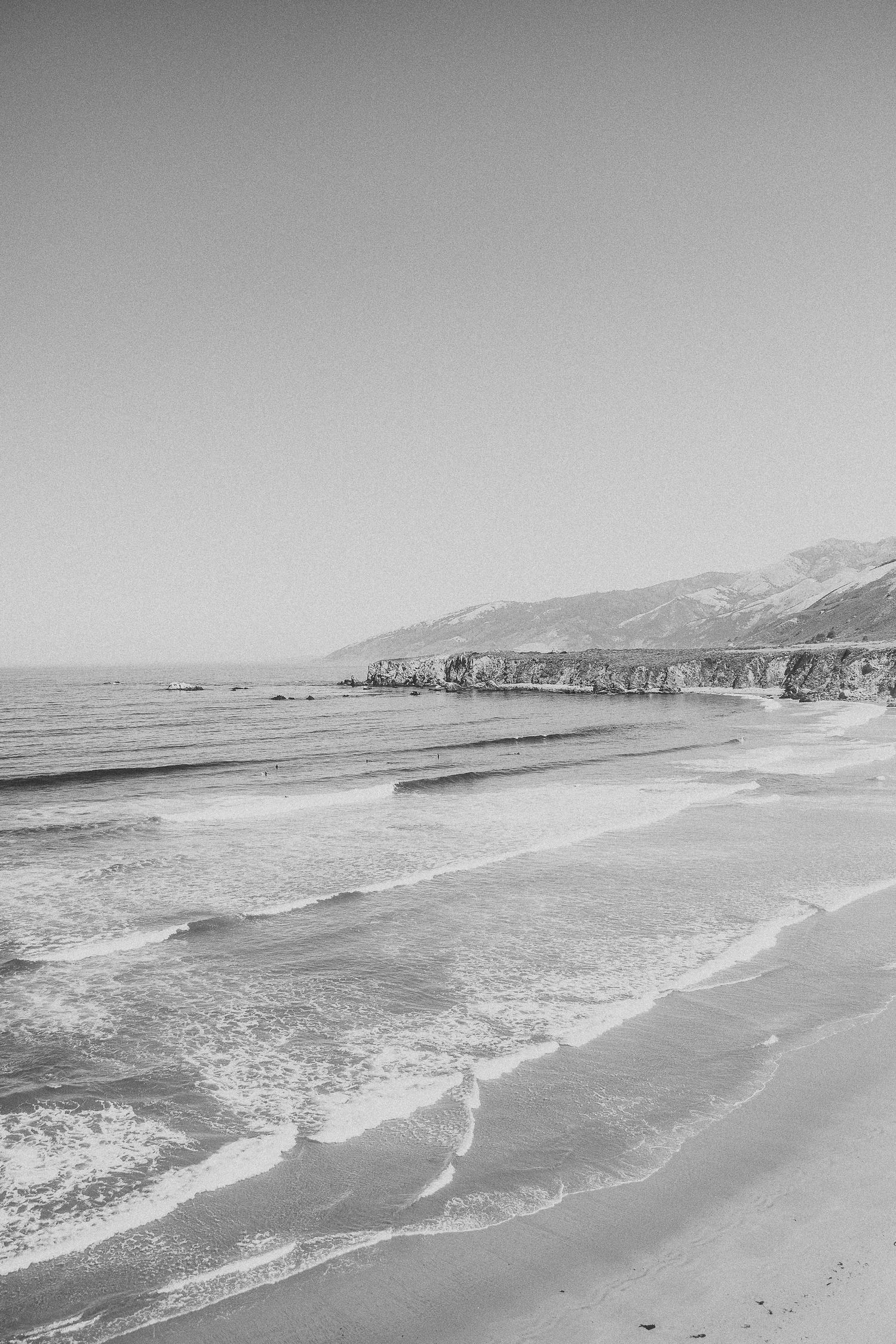 Black and white coastal landscape showing waves breaking on a beach with cliffs and mountains in the distance.