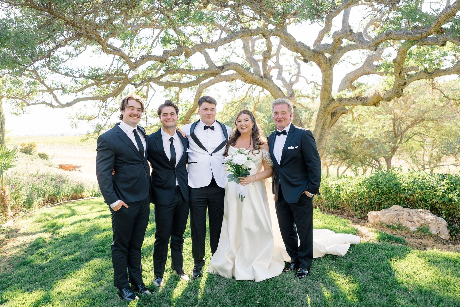 Bride in white gown poses with groom in tuxedo and three men in black suits under a sprawling oak tree.