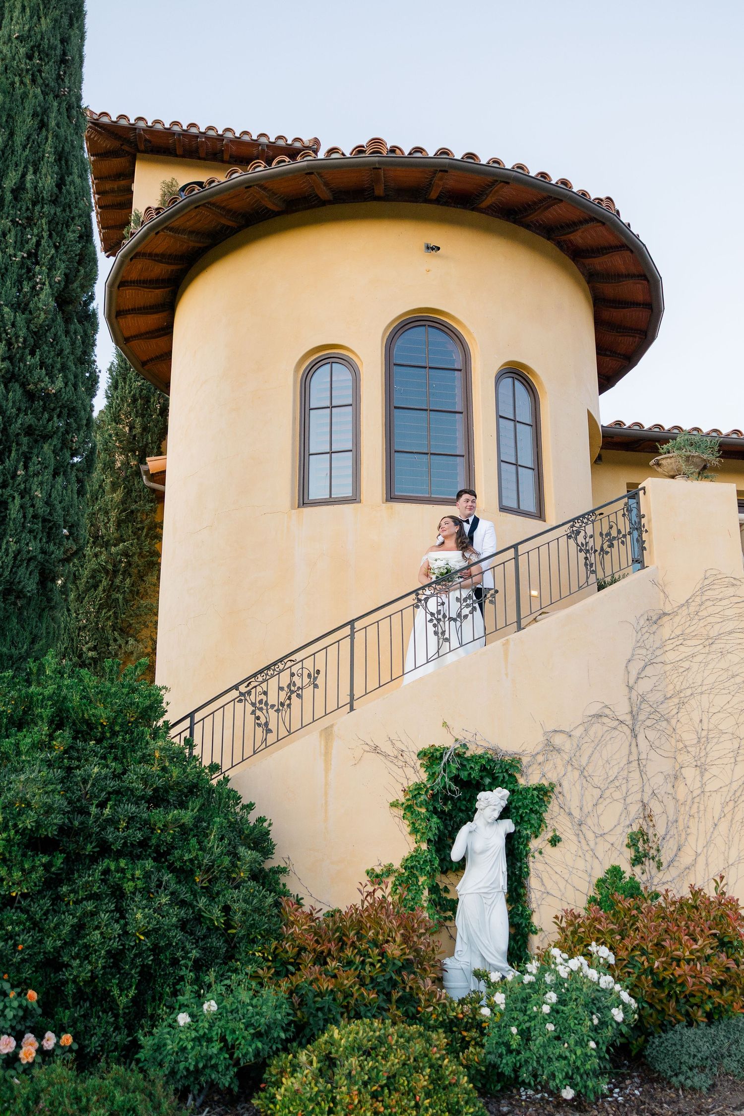 Bride and groom posing on ornate balcony of Tuscan-style villa surrounded by cypress trees.