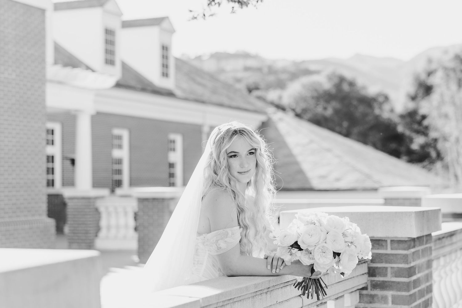 Bride in white gown and veil poses with bouquet on brick balcony in elegant black and white wedding portrait.