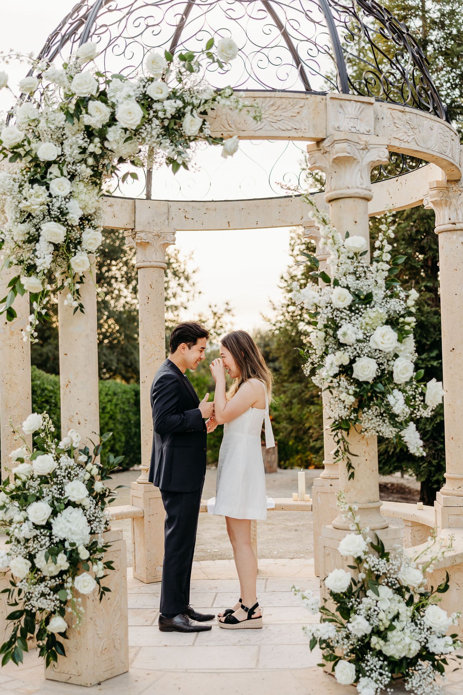 A romantic moment under an ornate white gazebo adorned with cascading white flowers and greenery.