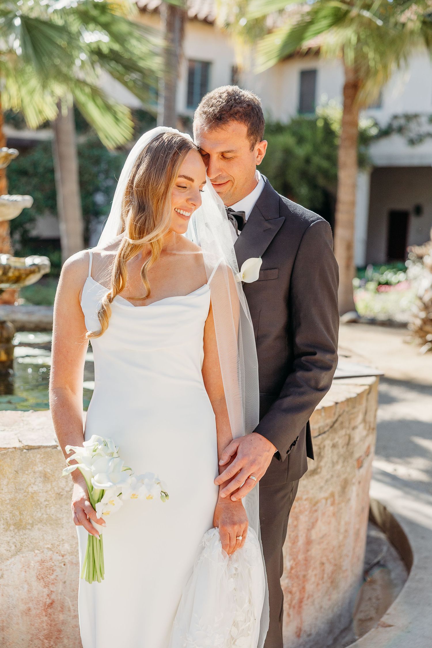 Newlyweds share intimate moment against tropical palm tree backdrop in elegant wedding attire.