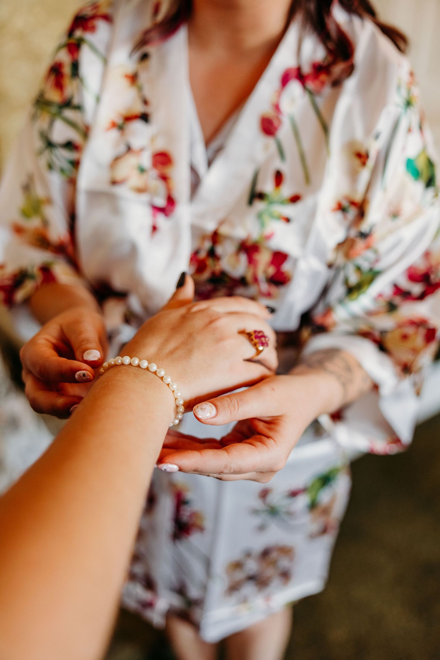 Hands placing delicate pearl bracelet on wrist with floral robe in background.