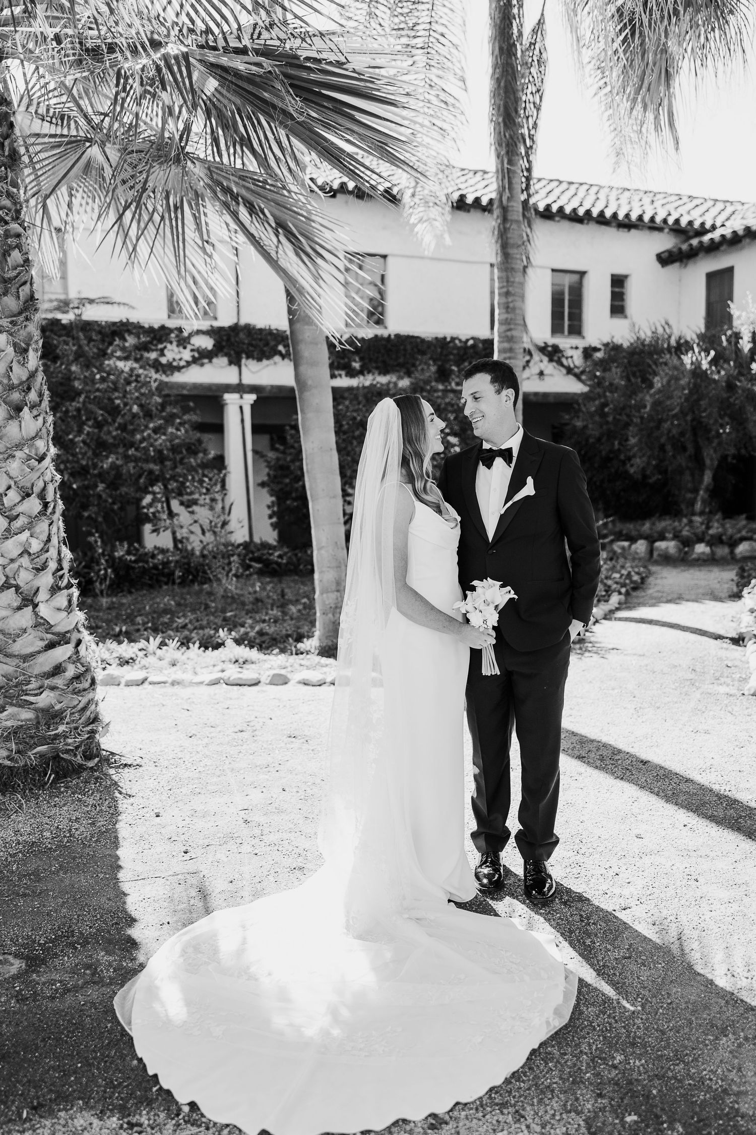 Black and white wedding photo of a couple standing in a garden with palm trees and Spanish-style architecture.