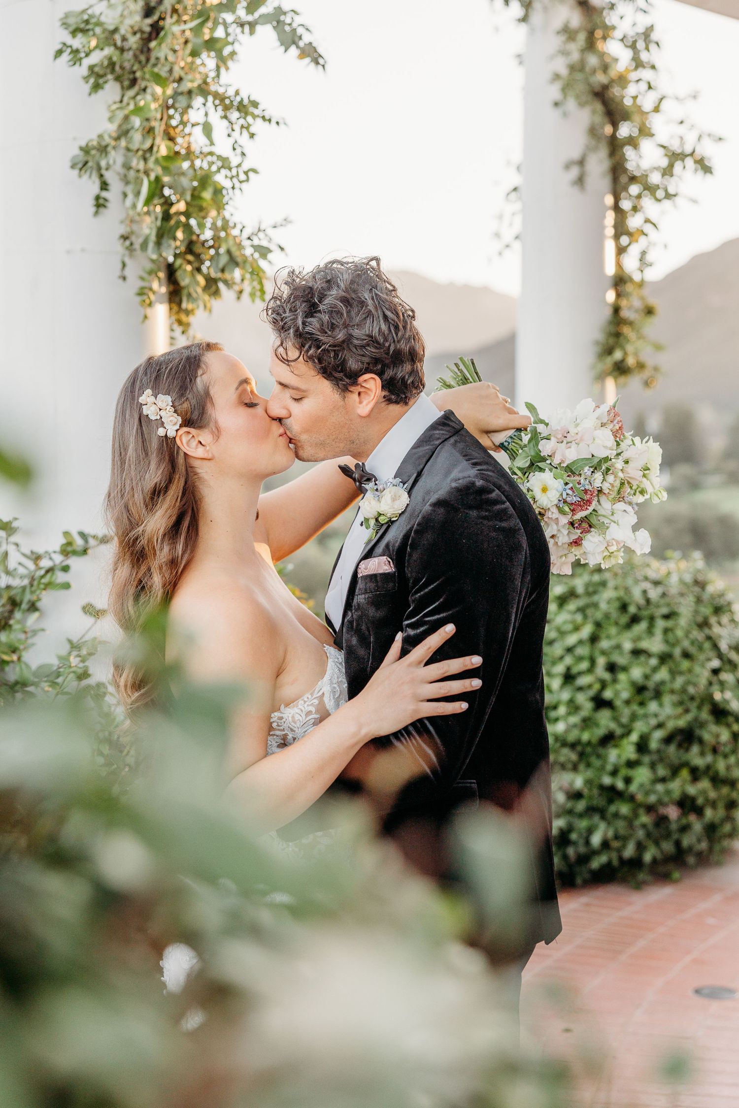 Romantic wedding moment captured under a floral-adorned pergola with greenery and natural light.