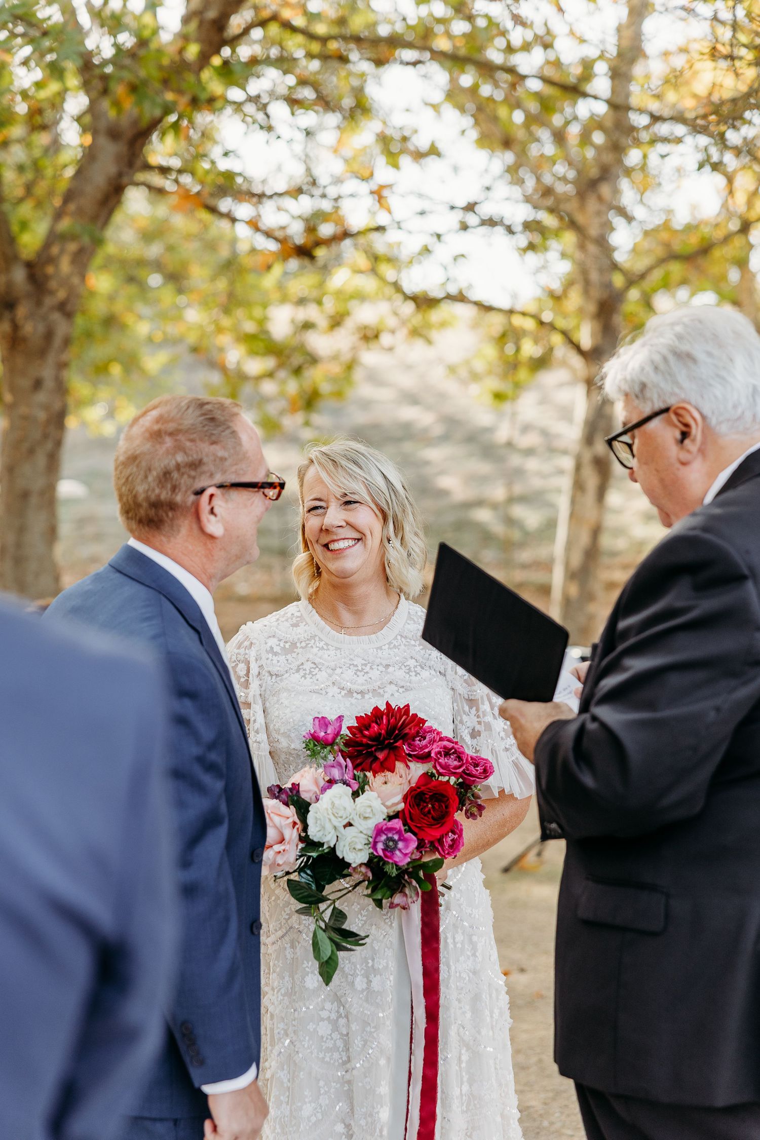 Wedding party gathered outdoors in autumn with bride holding vibrant red and white bouquet.
