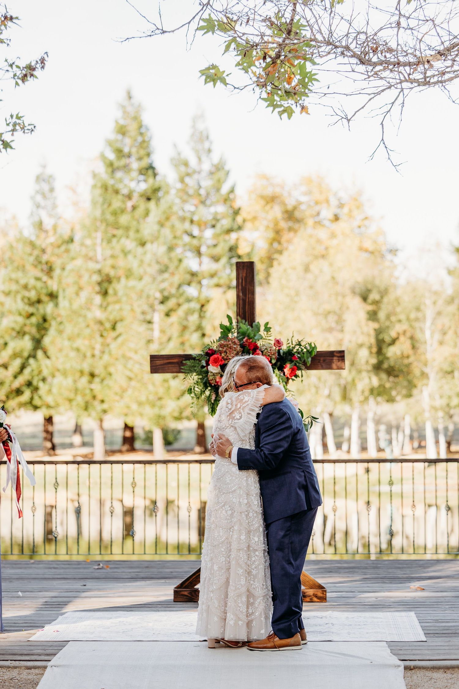 Newlyweds embrace during ceremony in front of wooden cross with evergreen trees in background.