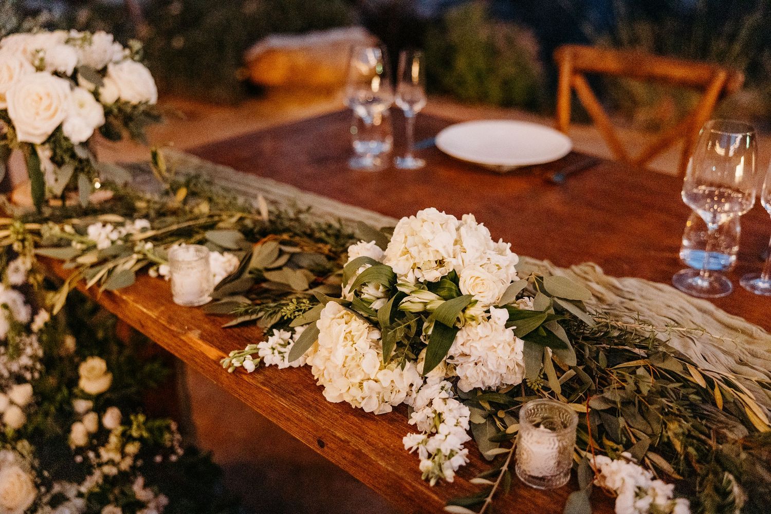 Elegant wedding reception table decorated with white floral arrangements and greenery garlands on wooden surfaces.