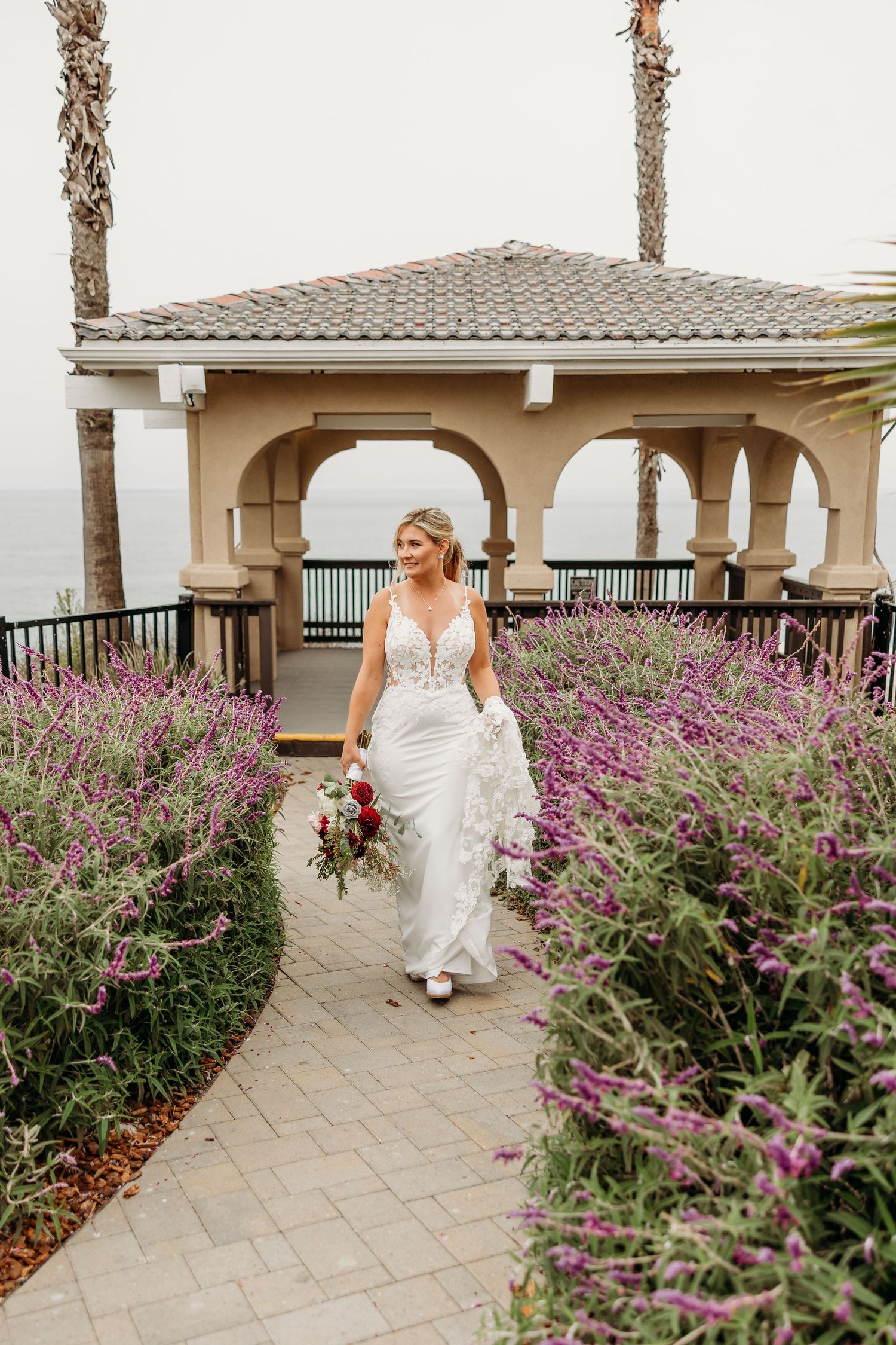 Beautiful wedding dress captured on a stone path with purple flowers and Mediterranean-style architecture.