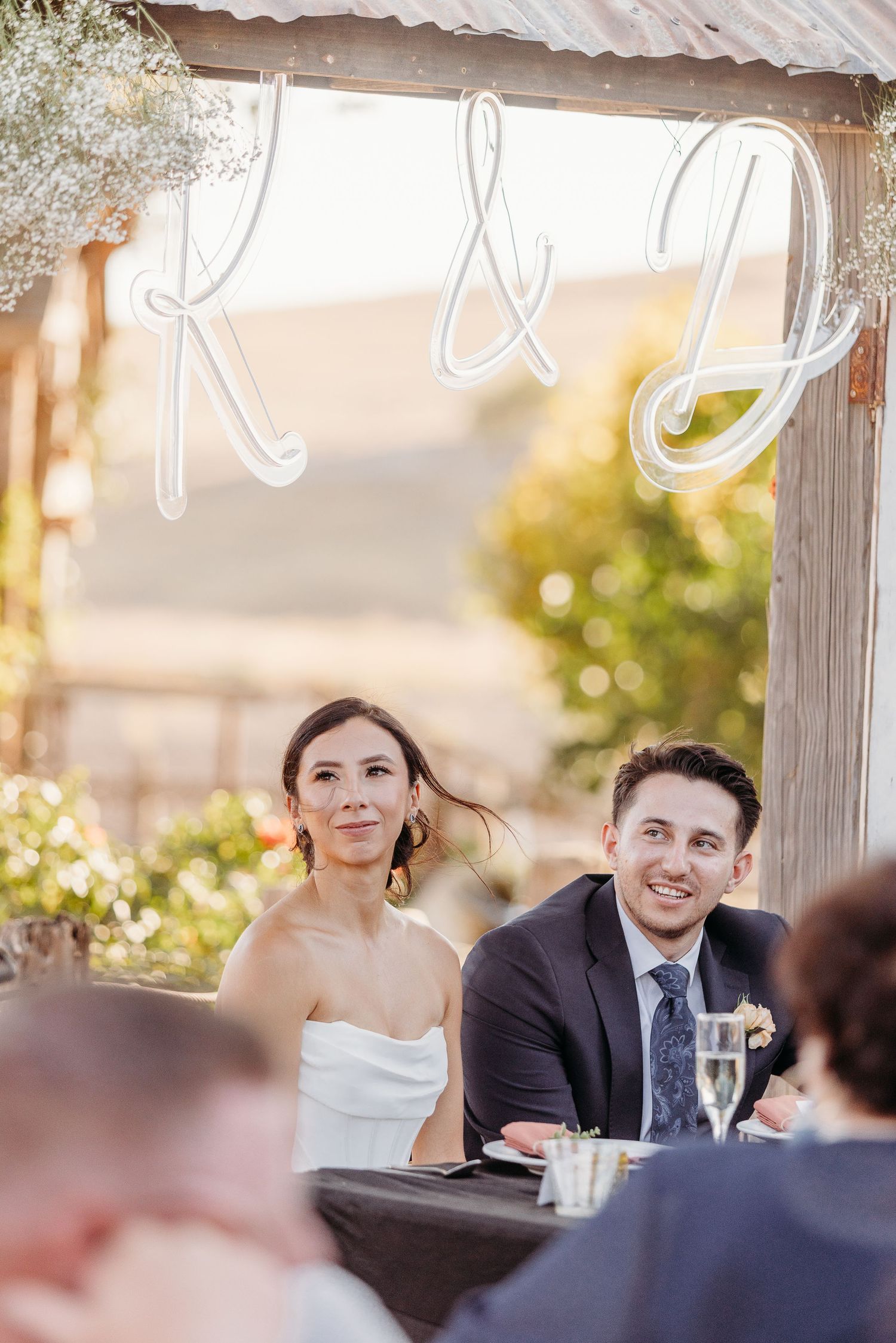 Two people seated at an outdoor dining table during sunset with a soft golden glow in the background.