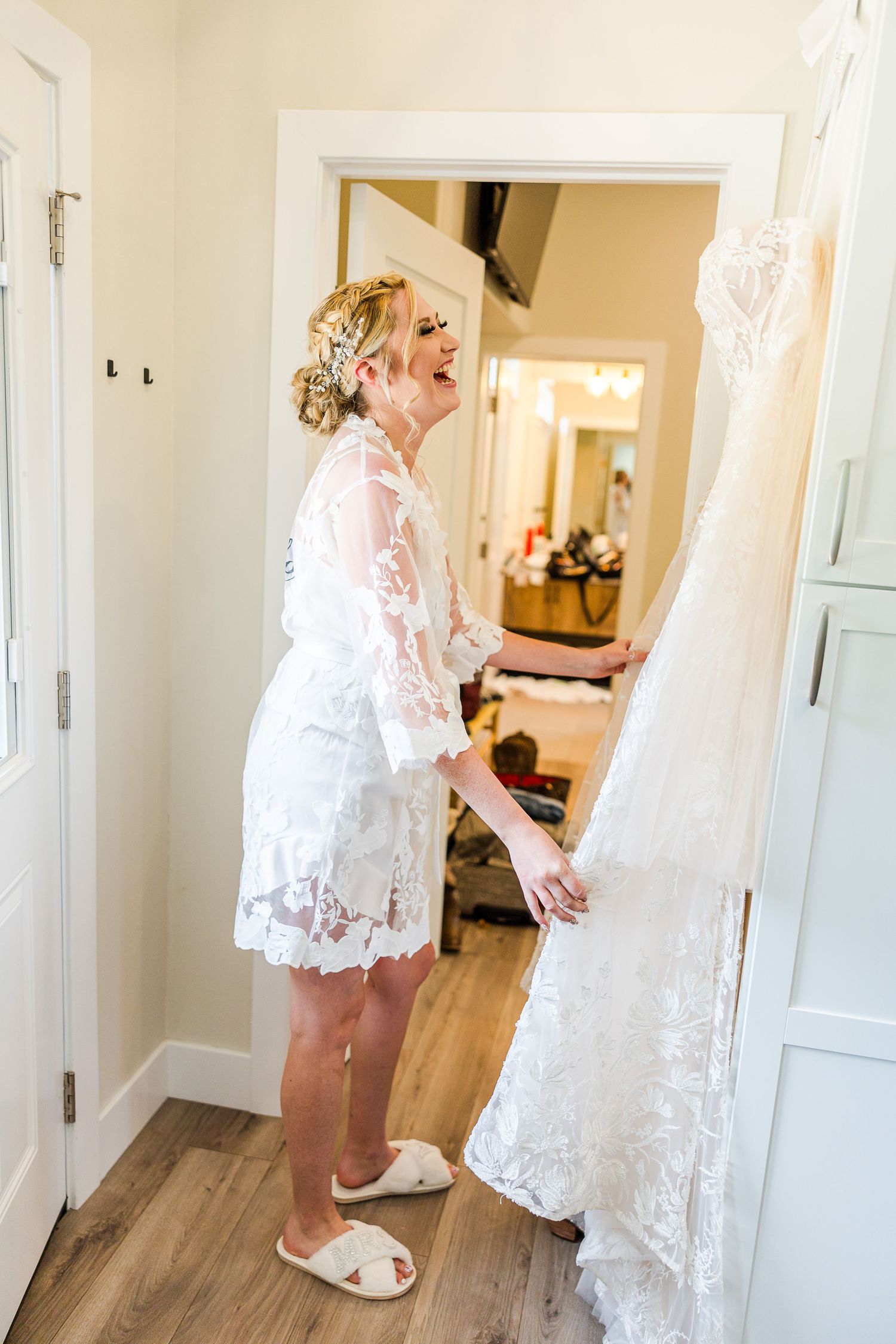A person in a white lace robe and slippers admires a wedding dress hanging in a doorway.