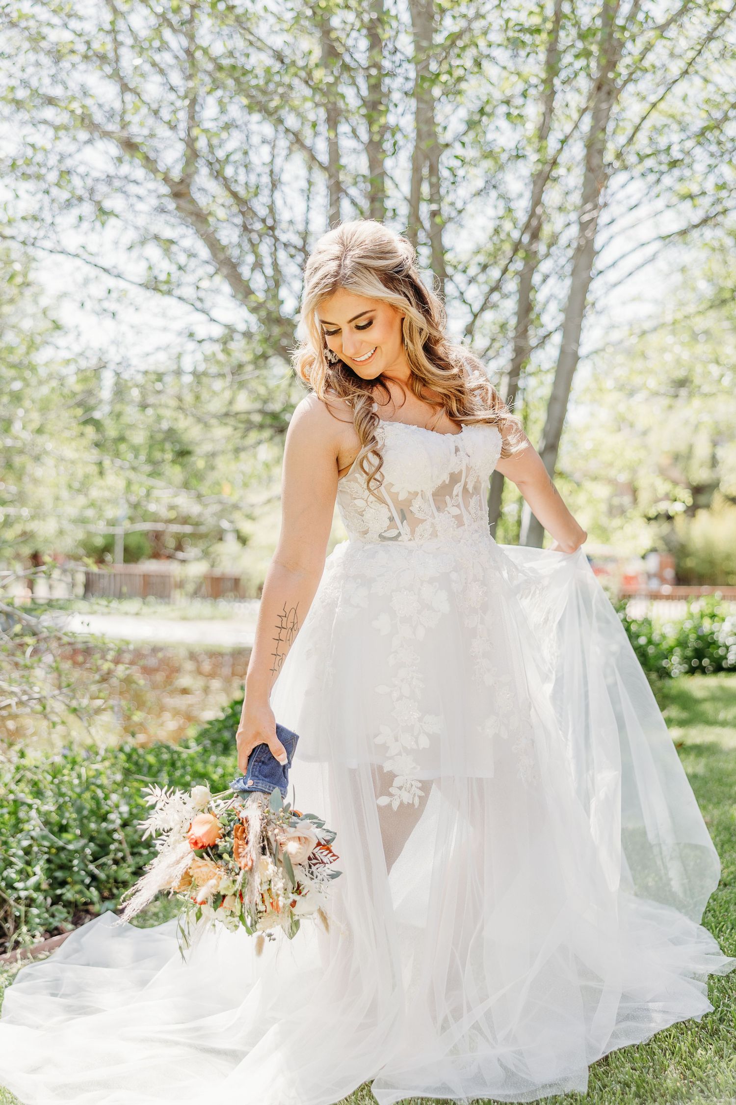 A bride in a flowing white lace gown holds a bouquet while standing in a sunlit garden setting with trees in background.
