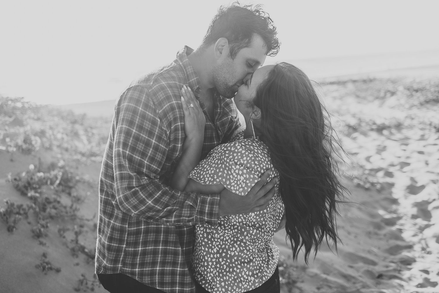 Black and white photo of couple embracing on sandy dunes.