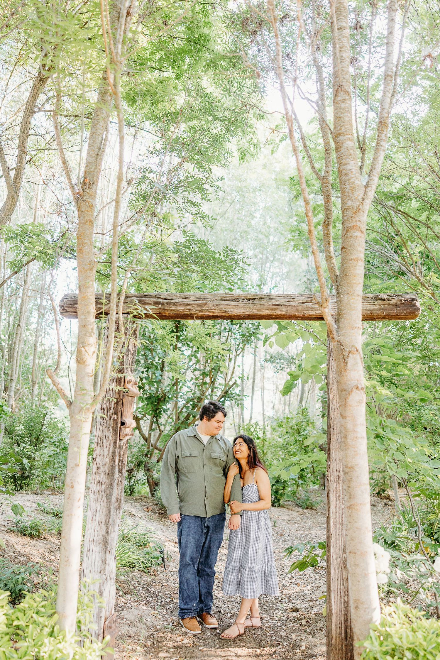 A couple sharing a romantic moment under a rustic wooden arch in a dreamy forest setting.