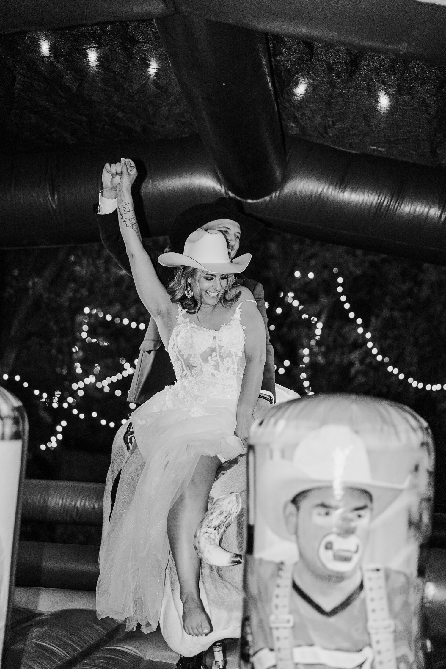 A bride in western attire rides a mechanical bull at a wedding reception.