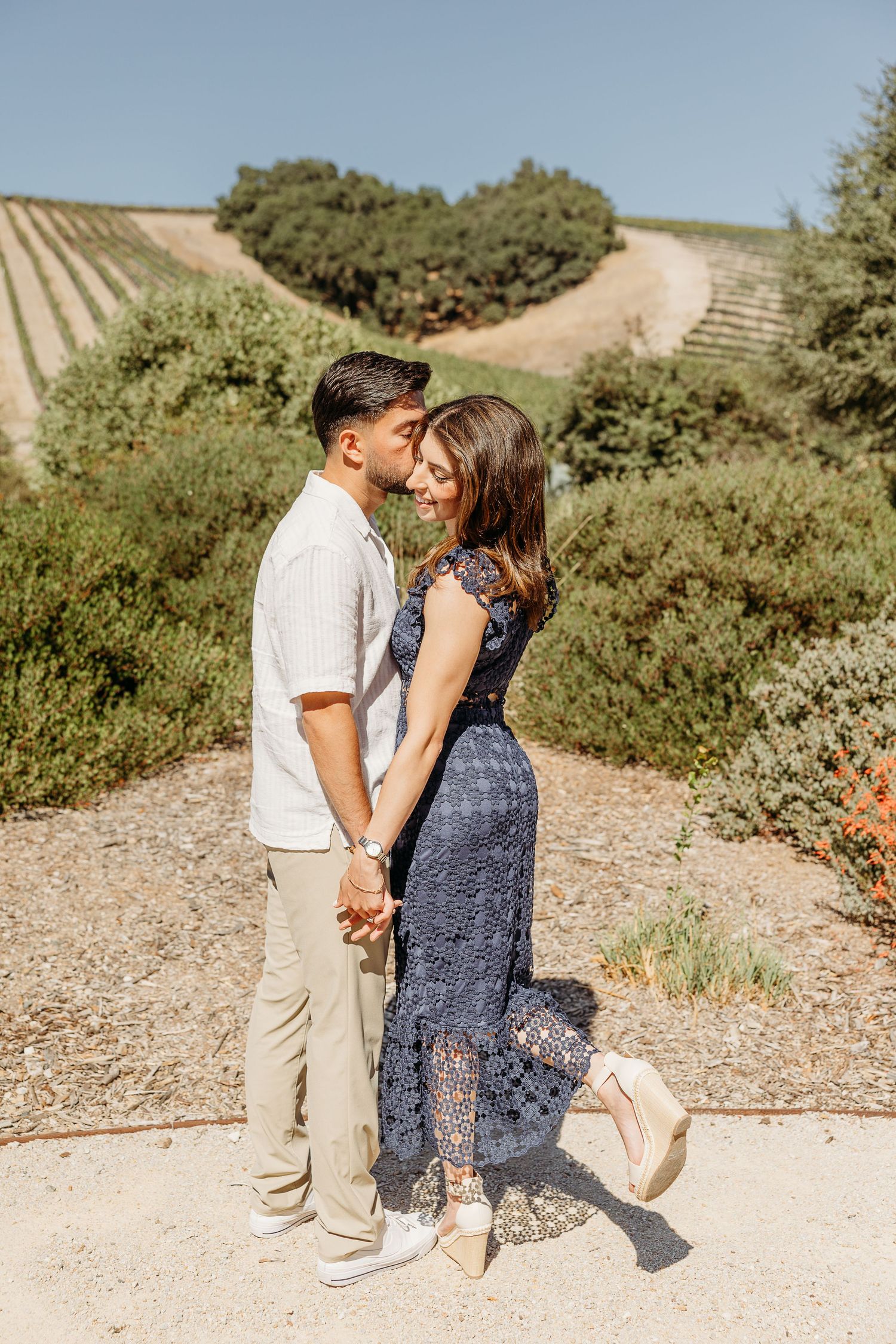 Couple shares intimate moment on dirt path surrounded by hillside vineyards.